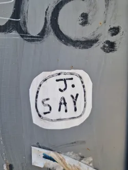 Street sticker A white sticker with black hand-drawn text that says J. SAY on a grey wall. The sticker has a slightly rounded square shape with a simple black border around the text.