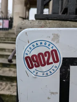 Street sticker A circular sticker with a white background and a prominent blue outer ring. Within this ring, the word 'BIDDANOBA' is written in blue capital letters along the top curve, and 'DE FORRU' is written in blue capital letters along the bottom curve. The center of the sticker features large, bold red numbers '09020'. The sticker appears slightly worn and has small water droplets visible on its surface, indicating it's a physical sticker affixed to a white, scratched, and somewhat dirty surface.