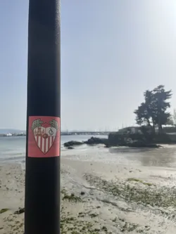 Street sticker A shield-shaped sticker featuring the official crest of Sevilla Fútbol Club. The crest is predominantly red and white, with a red border. At the top, two green laurel wreaths frame the shield. The upper left section of the shield displays three figures in a golden-brown tone, while the upper right section features a stylized 'SFC' monogram in white on a black background, with a brown football beneath it. The lower half of the shield is composed of vertical red and white stripes. The entire crest is set against a rectangular red background, which shows slight fading and minor wear, indicating it's a physical sticker.