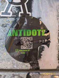 Street sticker A round black and green sticker with the word 'ANTIDOTE' in large green letters. Below the text, there is a QR code and Instagram handle @antidote_26. The sticker is slightly damaged and peeling from the surface. It is attached to a wall with other graffiti and stickers.