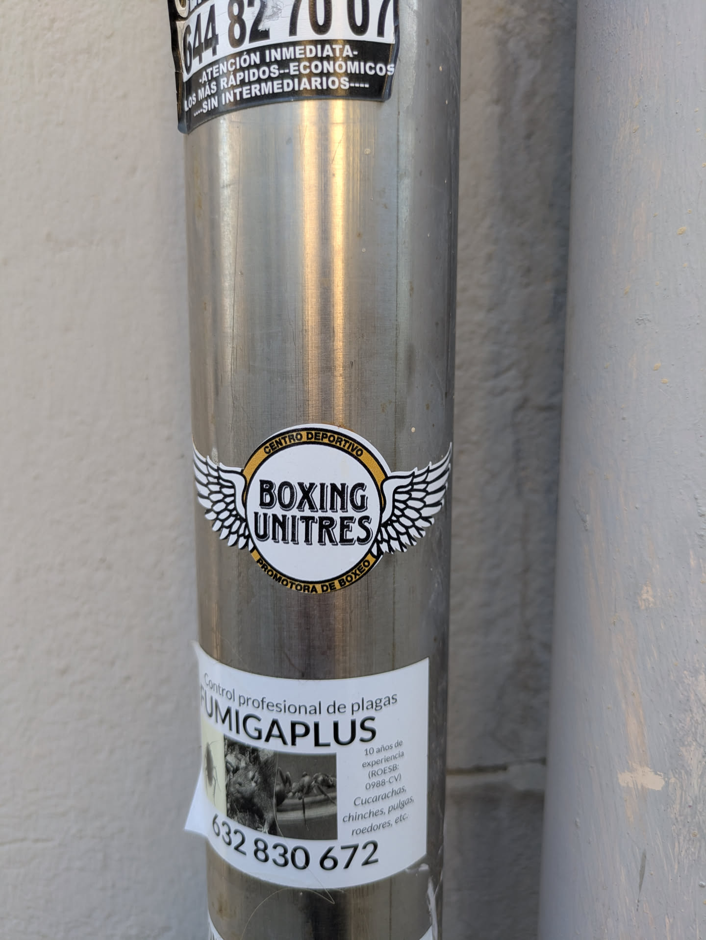 This image shows a close-up of a silver metal pole with two stickers on it. The upper sticker features the logo of a boxing club. It has the text 'CENTRO DEPORTIVO BOXING UNITRES PROMOTORA DE BOXEO' in a circular arrangement and also contains stylized wings. Below it, the second sticker advertises a pest control service called 'FUMIGAPLUS', mentioning 10 years of experience and listing pests controlled such as cockroaches, bedbugs, fleas and rodents. A phone number, '632 830 672', is prominently displayed.