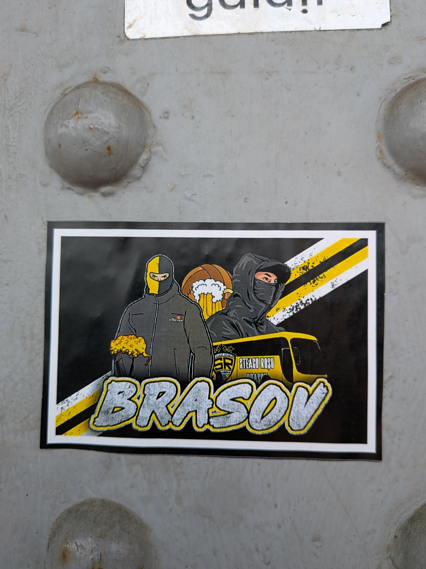Rectangular sticker featuring two masked football supporters, a beer mug, diagonal yellow and black stripes, and a yellow bus. The bottom displays 'BRASOV' in large, stylized grey letters.