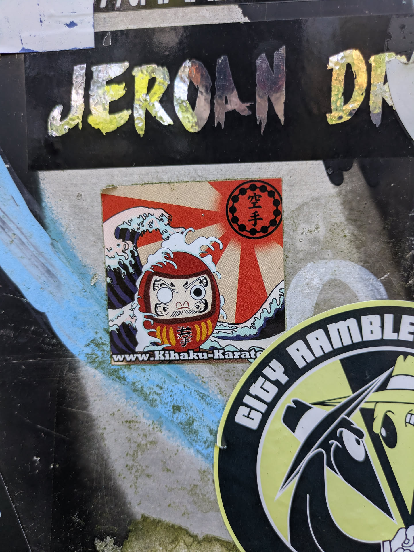 The central sticker depicts a red Daruma doll with one eye filled in, set against a dynamic background of stylized blue and white ocean waves and a prominent red rising sun (Hinomaru). Above the Daruma, within the sun's rays, the Japanese characters for 'Karate' (空手) are displayed inside a black circular border. On the Daruma's body, another Japanese character, '拳' (ken, meaning fist), is visible. Along the bottom edge of the sticker, a website address 'www.Kihaku-Karate.com' is printed.
