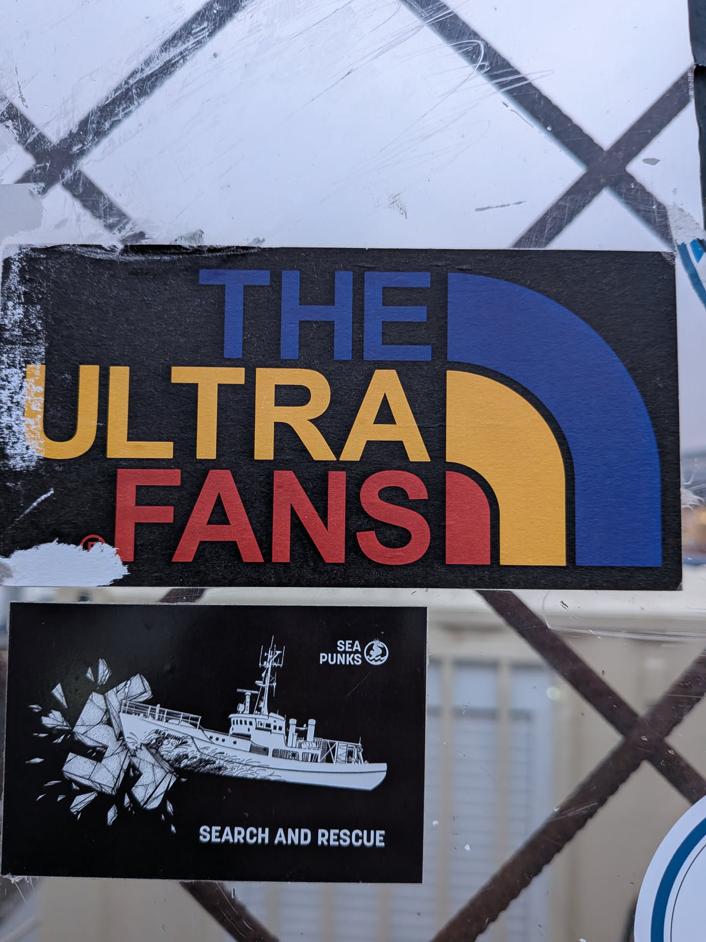 Street sticker The sticker is rectangular with a black background. It features large, bold text in three rows. The top row reads 'THE' in blue. The middle row reads 'ULTRA' in yellow. The bottom row reads 'FANS' in red. To the right of the text, a stylized semi-circle design is present, mimicking the iconic North Face logo, with a blue outer arc and a yellow inner arc. The sticker appears to be slightly worn at the edges.