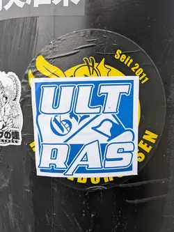 Street sticker A rectangular sticker with slightly rounded corners, predominantly blue and white. It features the word 'ULTRAS' in large, stylized block letters. The letters are white with a thick blue outline, set against a blue background. Intricate, abstract designs are visible within some of the letters, particularly the 'L' and 'R', adding to its dynamic appearance. The sticker shows signs of wear, including minor scuffs and slight peeling, indicating its physical nature. It is centrally placed over a larger, circular black and yellow sticker.