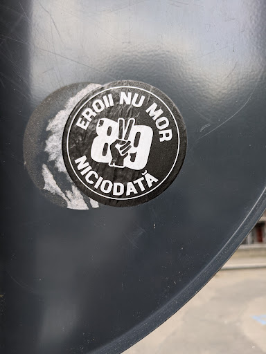 The sticker is on a grey surface. The black sticker has white text around the outer edge and a white peace sign hand gesture over the number 89 in the center. The text reads "EROII NU MOR NICIODATĂ", which translates to "Heroes never die". The sticker appears to be a memorial of the Romanian Revolution of 1989.