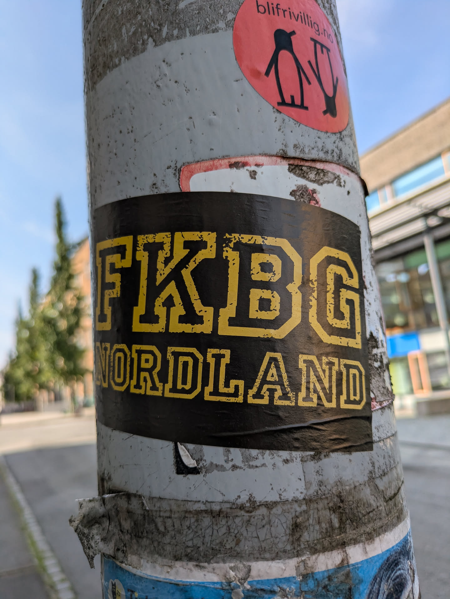 The central sticker is rectangular, featuring bold, distressed yellow block letters on a black background. The top line reads 'FKBG' and the bottom line reads 'NORDLAND'. The sticker is applied to a heavily textured, light-colored pole, which also shows signs of other old, peeling stickers underneath and above it. The overall impression is one of urban marking or tagging in a public space.