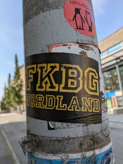 Street sticker The central sticker is rectangular, featuring bold, distressed yellow block letters on a black background. The top line reads 'FKBG' and the bottom line reads 'NORDLAND'. The sticker is applied to a heavily textured, light-colored pole, which also shows signs of other old, peeling stickers underneath and above it. The overall impression is one of urban marking or tagging in a public space.