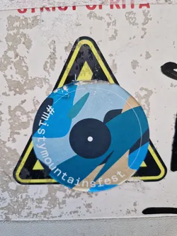 Round sticker with a design featuring abstract shapes in shades of blue and beige, resembling a stylized record.  Text on the sticker reads '#MistyMountainsFest'. The sticker is placed on top of a yellow and black warning triangle sticker.
