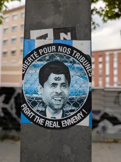 A round sticker on a pole features a caricature of Nasser Al-Khelaifi, president of Paris Saint-Germain, within a barbed wire circle. The text "Liberté pour nos tribunes" (Freedom for our stands) curves along the top, while "Fight the real enemy!" is along the bottom. The sticker is blue and white, with "QSI" (Qatar Sports Investments) marked on the caricature's forehead.