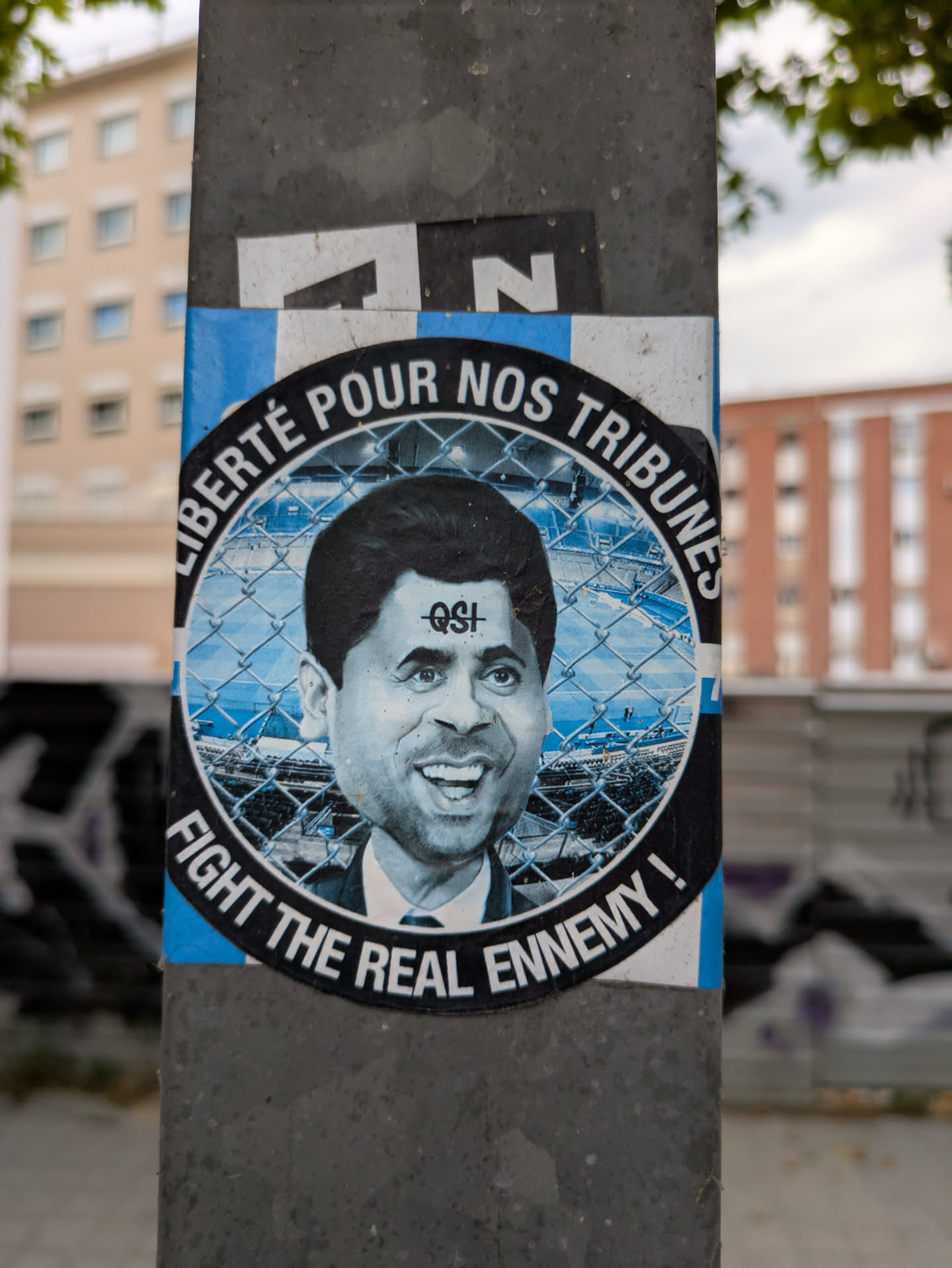 Street sticker A round sticker on a pole features a caricature of Nasser Al-Khelaifi, president of Paris Saint-Germain, within a barbed wire circle. The text "Liberté pour nos tribunes" (Freedom for our stands) curves along the top, while "Fight the real enemy!" is along the bottom. The sticker is blue and white, with "QSI" (Qatar Sports Investments) marked on the caricature's forehead.