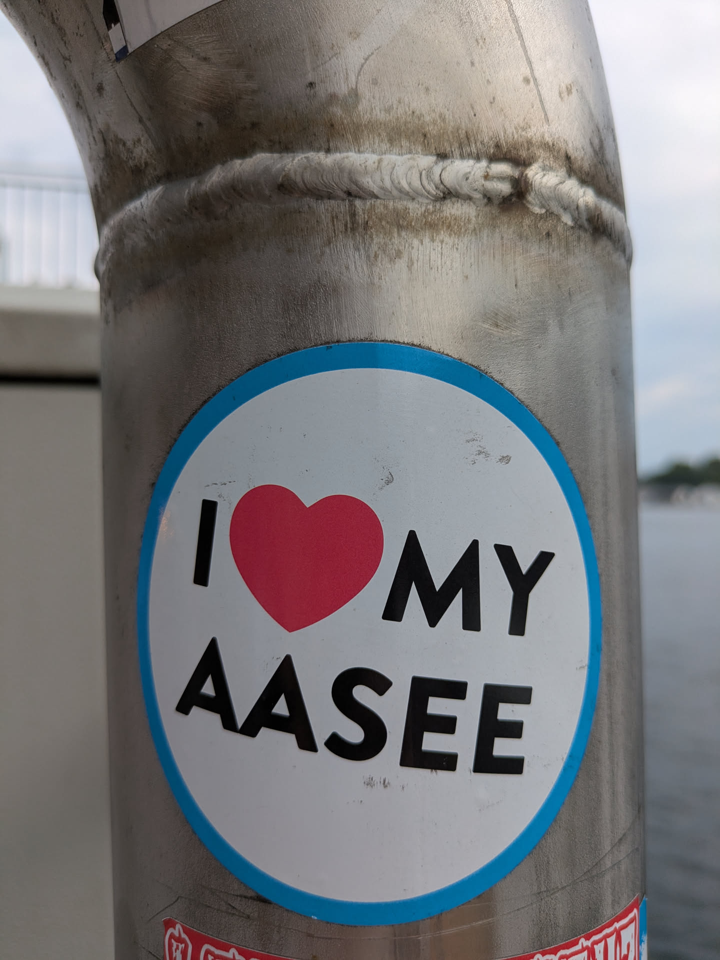 A white circular sticker with a light blue border, featuring the text 'I ❤️ MY AASEE'. The 'I' and 'MY AASEE' are in black capital letters, and a red heart symbol replaces the word 'LOVE'. The sticker is placed on a brushed or textured metal pole, which appears to be outdoors with water visible in the blurry background.