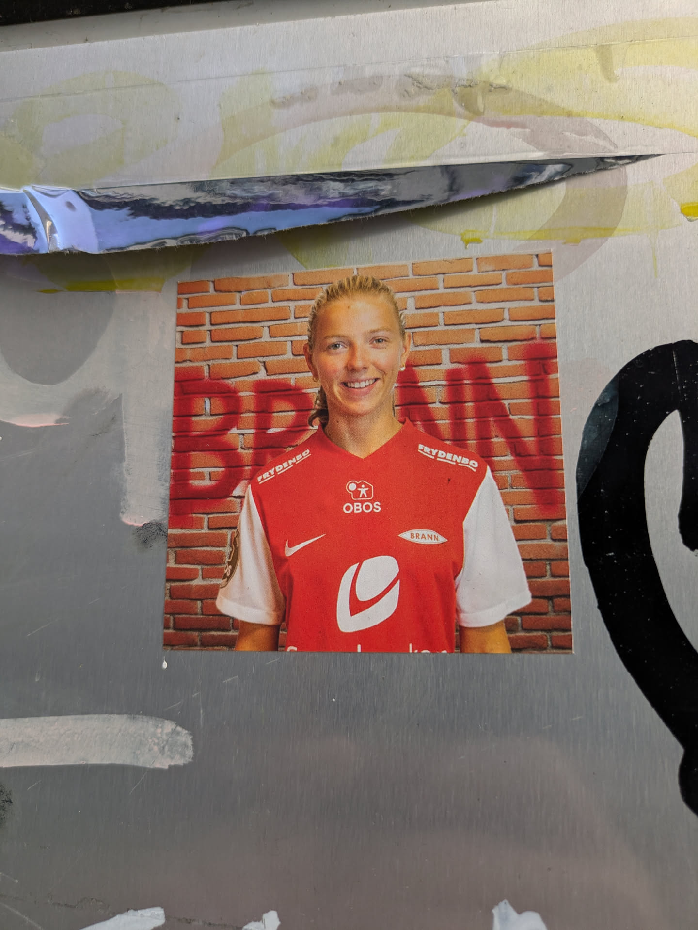 The sticker prominently displays a headshot of a fair-haired woman with a pleasant smile, wearing a red and white sports jersey. Her hair is pulled back, and she appears to be an athlete. The jersey features several sponsor logos, including 'FRYDENDO' on the shoulder, 'OBOS' and 'BRANN' on the chest, and a large white stylized 'L' or 'C' logo below 'BRANN'. The background behind her is a brick wall, with faded red graffiti that appears to spell 'BRANN' horizontally across the bricks. The overall impression is that of a professional sports portrait, likely a player for the SK Brann team.