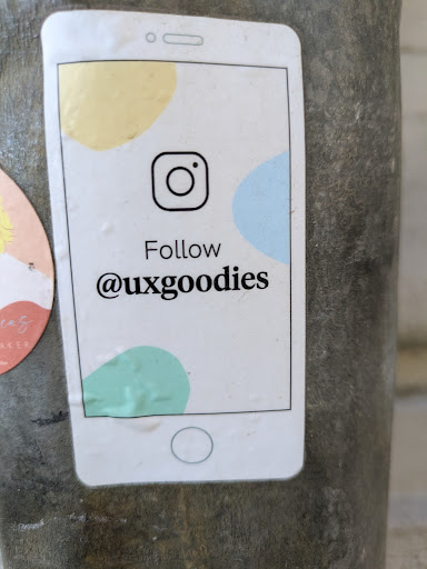 A sticker shaped like a white smartphone with a screen displaying the Instagram logo and the text 'Follow @uxgoodies'. The background has abstract shapes in yellow, blue, and green.