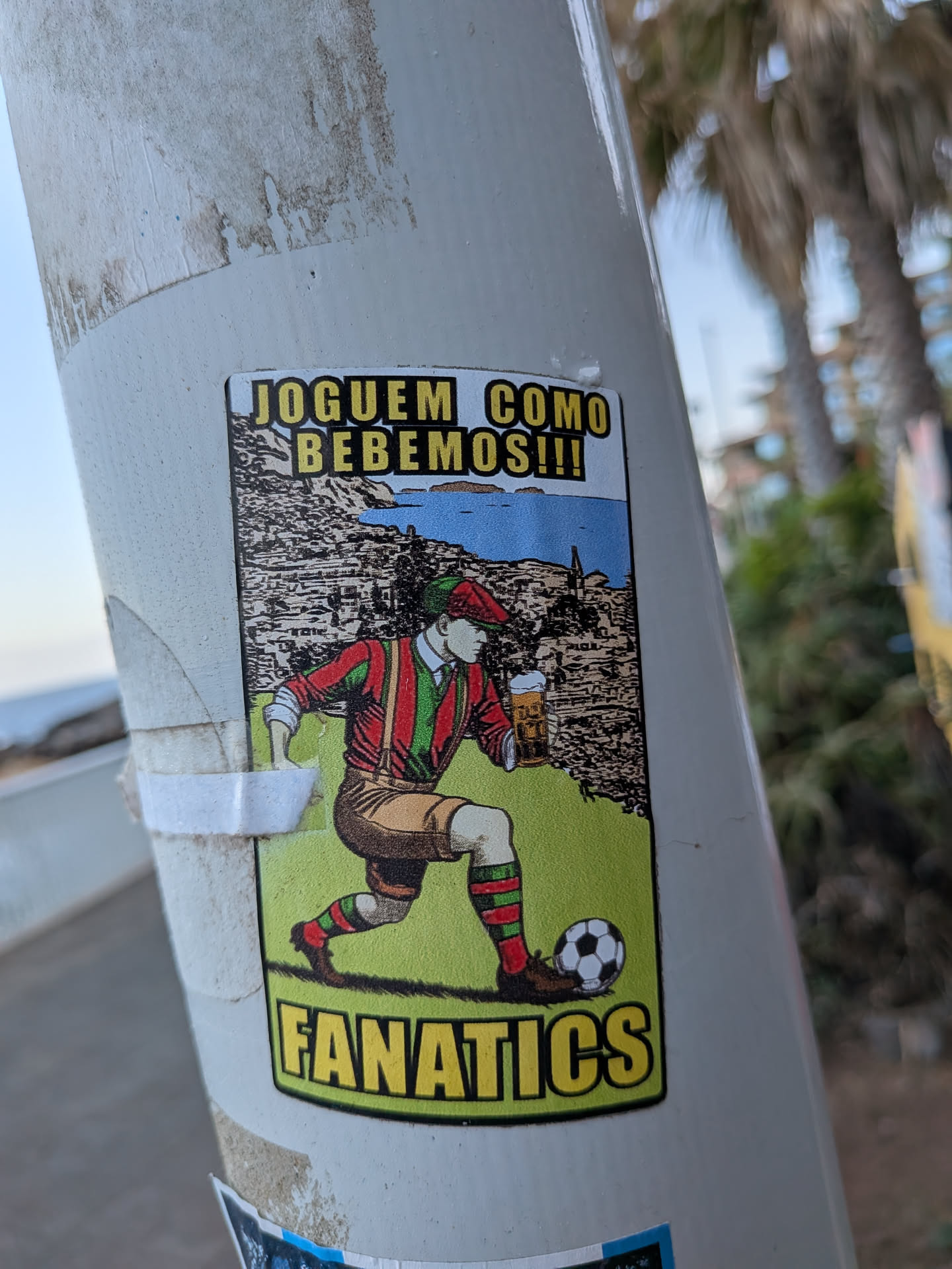 A rectangular sticker depicting a vintage-style soccer player in red and green gear dribbling a ball while holding a beer mug. Set against a coastal town background, it features yellow text reading 'JOGUEM COMO BEBEMOS!!!' at the top and 'FANATICS' at the bottom.