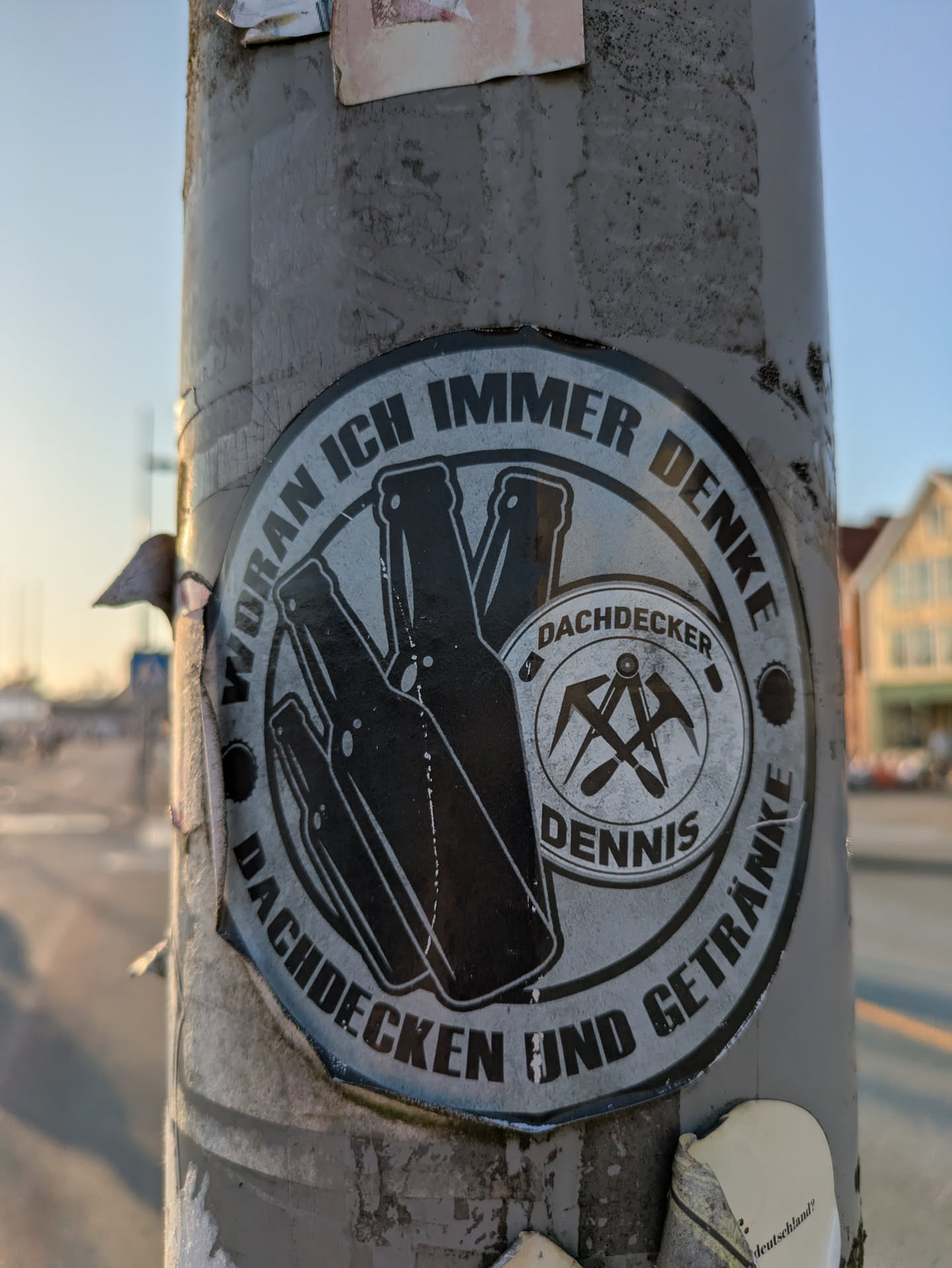 A weathered, circular black and white sticker is affixed to a grey metal pole. The sticker's design features a central inner circle containing the text 'DACHDECKER DENNIS' and a graphic of crossed roofing tools (likely a roofer's hammer and pick). Surrounding this inner circle, the main part of the sticker shows silhouettes of three beer bottles on the left, and a circular text that reads 'WORAN ICH IMMER DENKE' at the top and 'DACHDECKEN UND GETRÄNKE' at the bottom. The sticker appears to be peeling slightly at the edges.