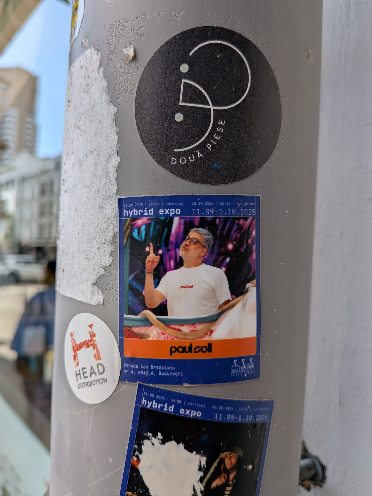 Rectangular sticker with a blue and orange design. It features a photo of a man in a white t-shirt pointing upwards, set against a colorful background. Text details an exhibition called 'hybrid expo' by 'paul soll' in Bucharest.