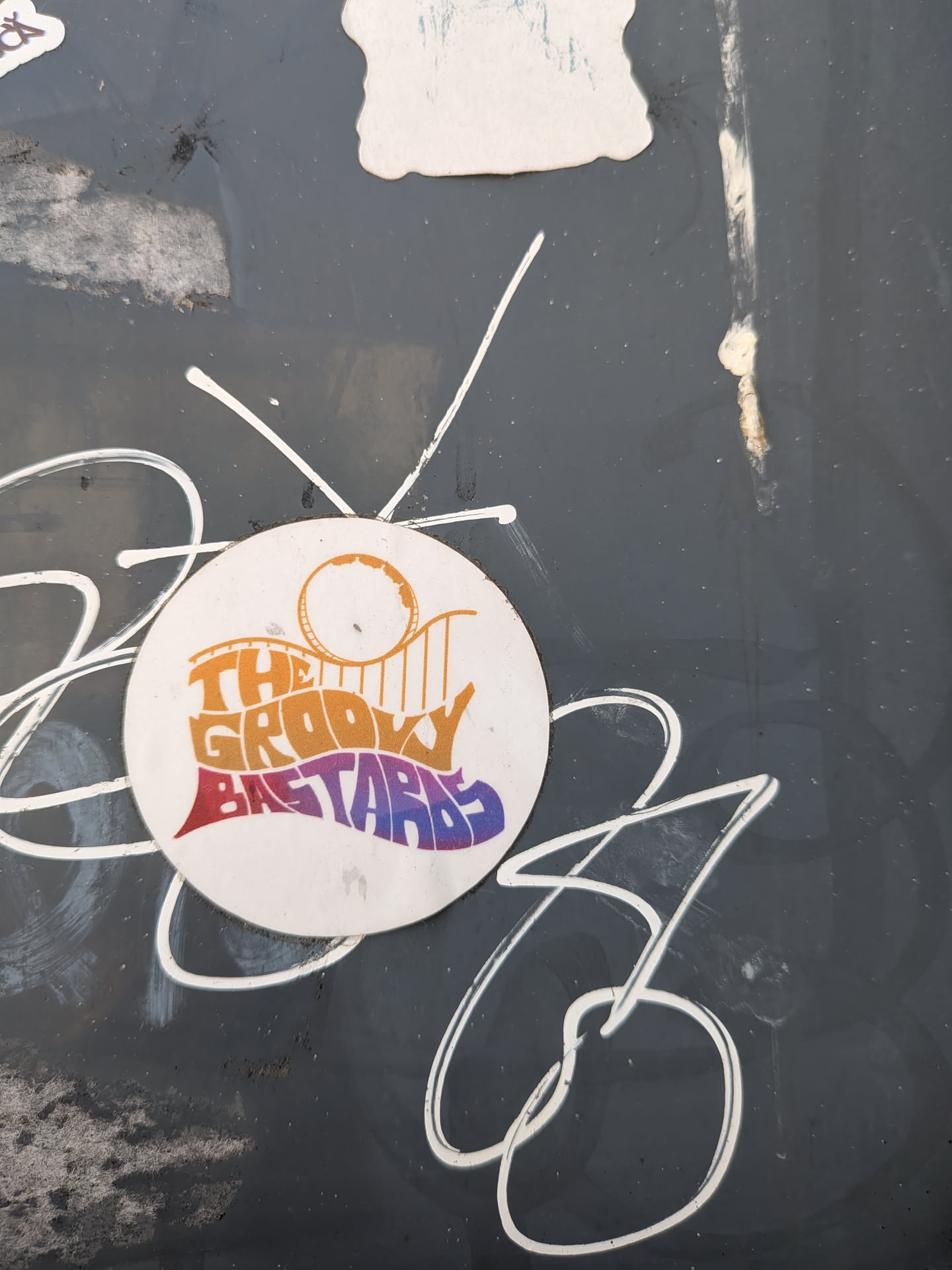 Circular white sticker featuring wavy, retro text 'THE GROOVY BASTARDS' in orange, red, and purple gradients. A minimalist orange roller coaster loop sits above the text.