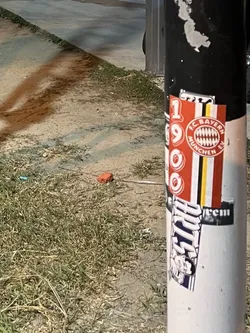The central sticker is a rectangular design for FC Bayern München. It features a vibrant red background on the left with large white numbers '1', '9', '0', '0' stacked vertically, likely representing the club's founding year. To the right of these numbers, the iconic circular FC Bayern München crest is prominently displayed, featuring a red and white checkered pattern within a red ring that reads 'FC BAYERN MÜNCHEN E.V.'. Further to the right, there are vertical stripes in red, yellow, black, and white. Below this main sticker, another sticker is partially visible. It's predominantly black and white/grey, featuring stylized text that appears to read 'FRITZ' and a cartoonish character's face with a mischievous expression, possibly a mascot or a brand character.