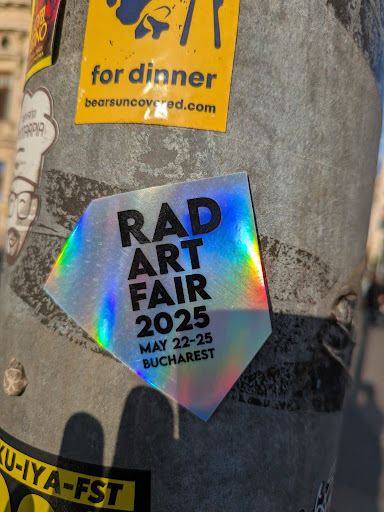 A holographic diamond-shaped sticker adhered to a pole promoting the RAD Art Fair 2025, taking place May 22-25 in Bucharest. The sticker is iridescent and catches the light, making the text "RAD ART FAIR 2025" stand out. Below the sticker there is another sticker.
