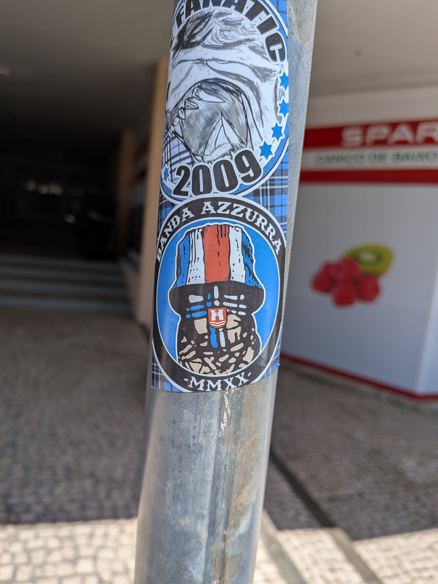 A vertical sticker with a blue plaid background wrapped around a metal post. It features a top graphic with the text 'FANATIC' and '2009', and a bottom circular emblem labeled 'BANDA AZZURRA' and '-MMXX-' depicting a figure in a blue, white, and red bucket hat.