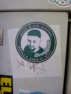 A square sticker featuring a circular dark green emblem. Inside is a stylized portrait of a smiling man. Italian text reads 'NON È UN ADDIO...' and 'BRILLA AMICO MIO!', with 'Bembono' in script.