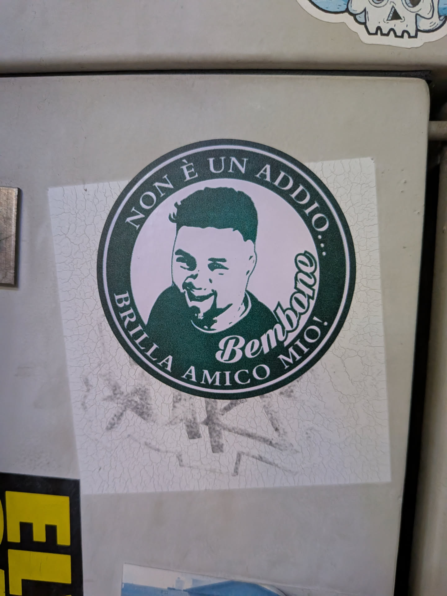 A square sticker featuring a circular dark green emblem. Inside is a stylized portrait of a smiling man. Italian text reads 'NON È UN ADDIO...' and 'BRILLA AMICO MIO!', with 'Bembono' in script.