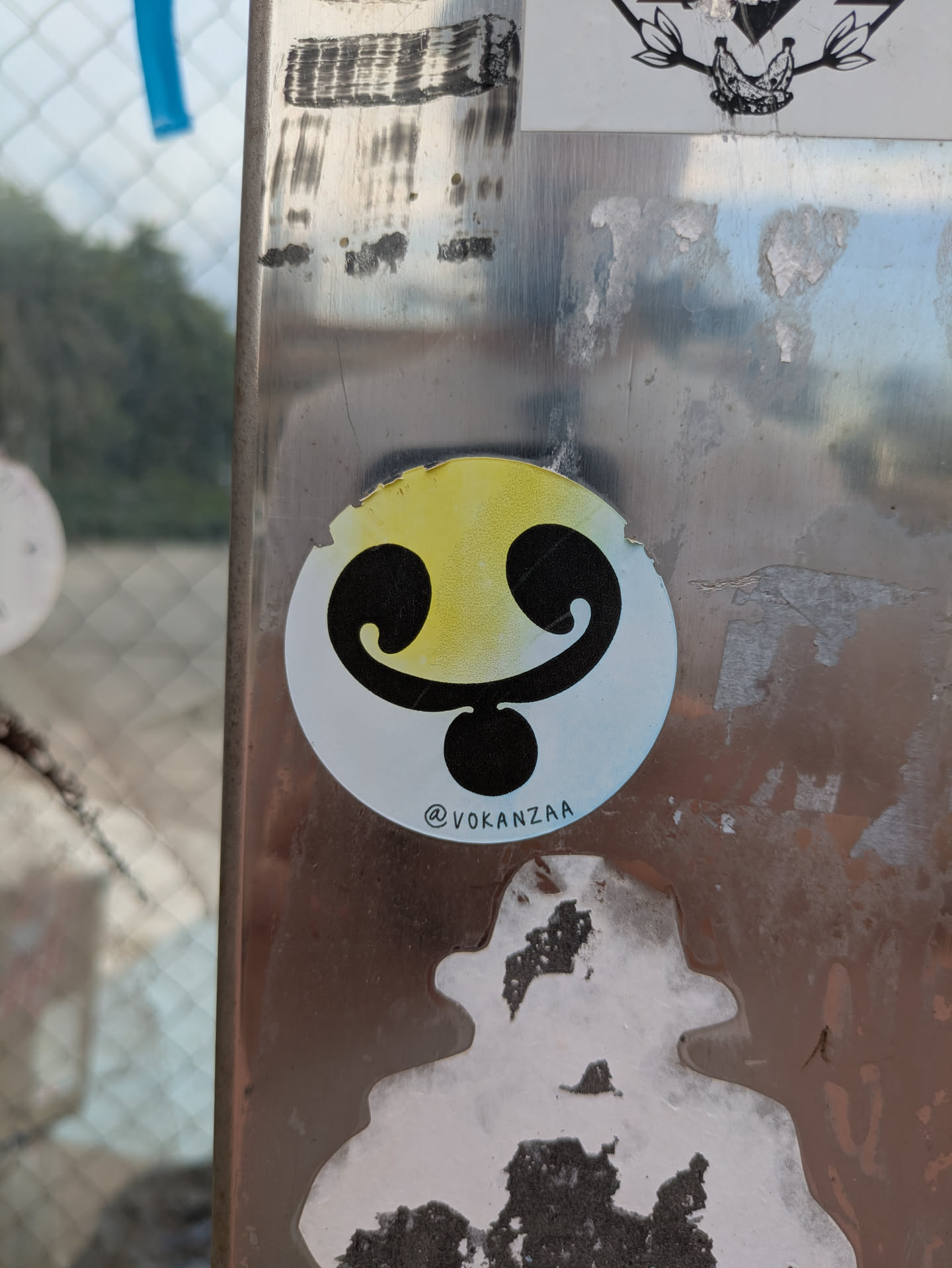 The sticker features a white background with a yellow gradient at the top. The design incorporates two curved black shapes resembling eyes or eyebrows, connected to a solid black circle that serves as the mouth or chin. The design is simple yet expressive. At the bottom of the sticker there is the text "@vokanzaa".