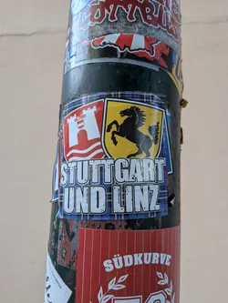 Street sticker Rectangular sticker with a blue tartan background. Features the coats of arms of Linz (red shield, white castle) and Stuttgart (yellow shield, black horse) above the bold white text 'STUTTGART UND LINZ'.
