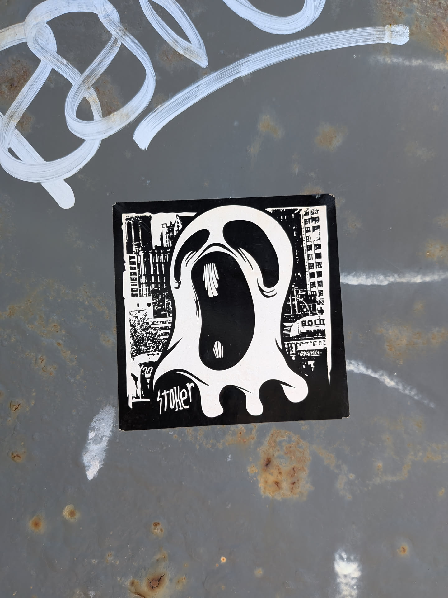 A square black and white sticker depicting a stylized, screaming white ghost. The background features a high-contrast urban cityscape. The word 'stoker' is written in stylized script at the bottom left.