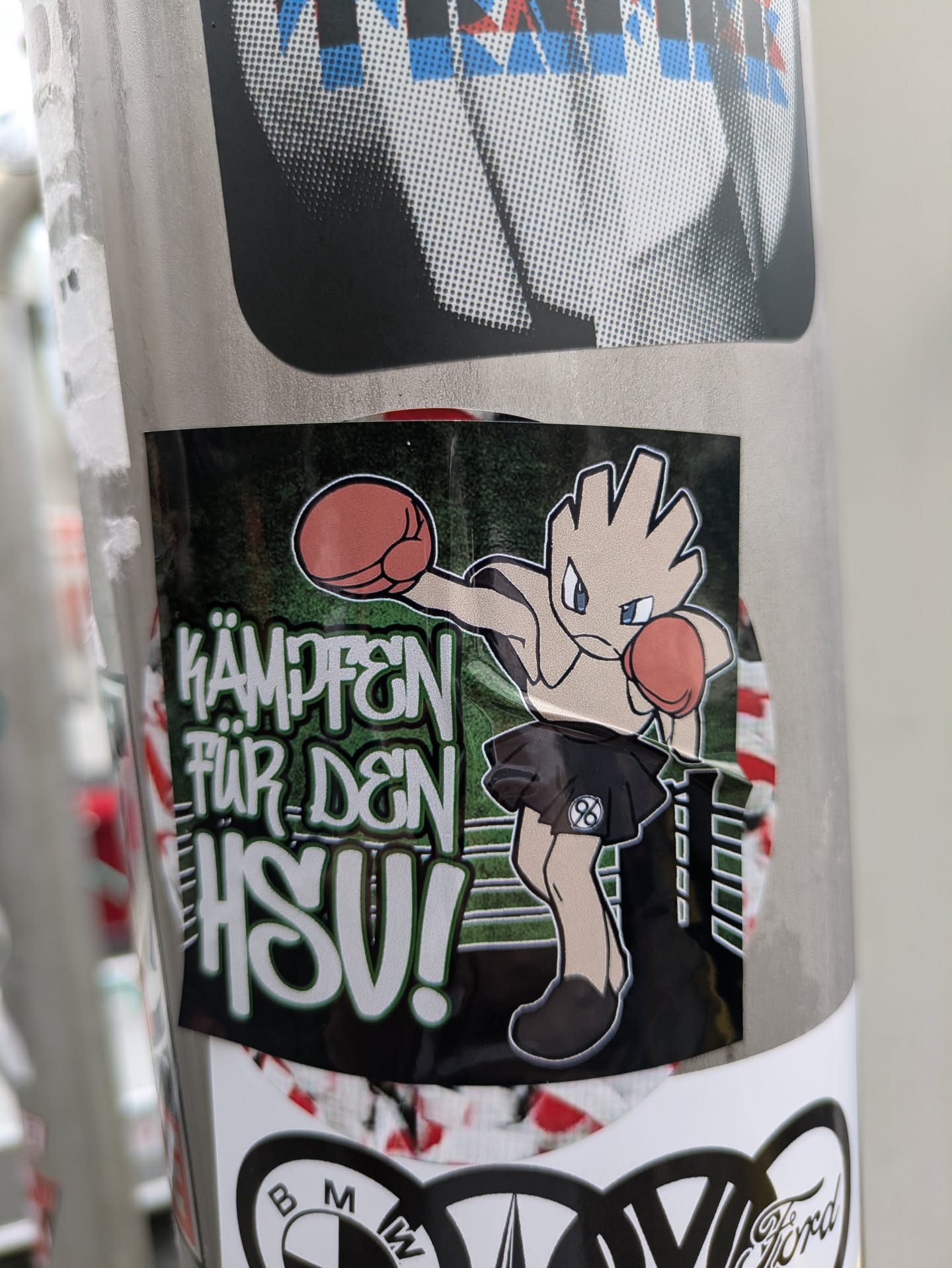 Street sticker The sticker prominently displays a stylized cartoon character resembling the Pokémon Hitmonchan. It has light beige skin, five-fingered hands, and blue eyes, wearing red boxing gloves and a black mini-skirt. On the skirt, a white '96' logo is visible within a circle. The character is depicted in a fighting stance, set against a dark green background with a subtle pattern that suggests a sports arena or field. Bold white text with a green outline reads 'KÄMPFEN FÜR DEN HSV!'. The overall design suggests support for a sports team, likely Hamburger SV (HSV), with a playful nod to pop culture and possibly another team like Hannover 96 due to the '96' logo.