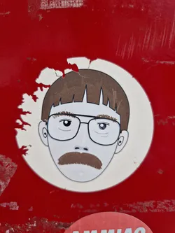 A sticker featuring an illustration of a man with glasses and a mustache. The style is simple and minimalist. The sticker is applied to a red surface that is slightly damaged.