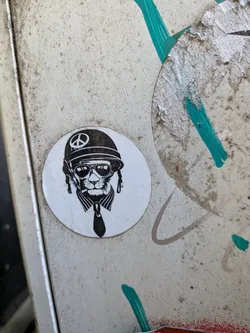 Street sticker Circular white sticker with a black ink drawing of a cat wearing a peace-sign helmet, sunglasses, and a tie.