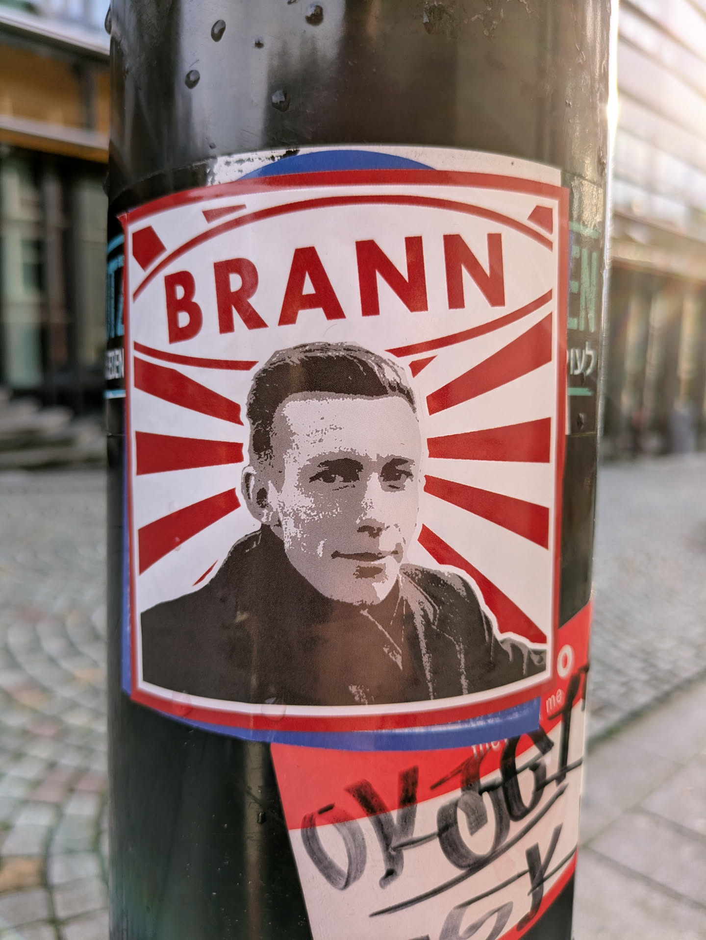 Street sticker The sticker, predominantly white, red, and black, displays a black and white, halftone-style portrait of a man with short, dark hair and a neutral expression, wearing a dark collar or jacket. Behind his head, red rays fan out against a white background, creating a sunburst effect. Above the portrait, the word "BRANN" is printed in large, bold red capital letters. The entire design is bordered by a thin red line, followed by an even thinner blue line. The sticker is affixed to a black, somewhat textured pole, which appears to have water droplets on its surface. Partially visible below this sticker are other obscured stickers or graffiti.