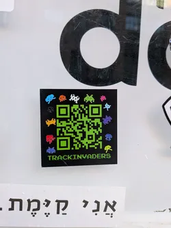 Street sticker A square-shaped sticker featuring a vibrant green QR code against a black background. Surrounding the QR code are numerous small, pixelated alien characters, reminiscent of the classic Space Invaders game, rendered in a variety of bright colors including blue, orange, white, green, pink, black, yellow, red, and purple. Below the QR code, the word 'TRACKINVADERS' is written in a pixelated green font, matching the QR code's color. The sticker has a glossy finish and is applied to a white surface.