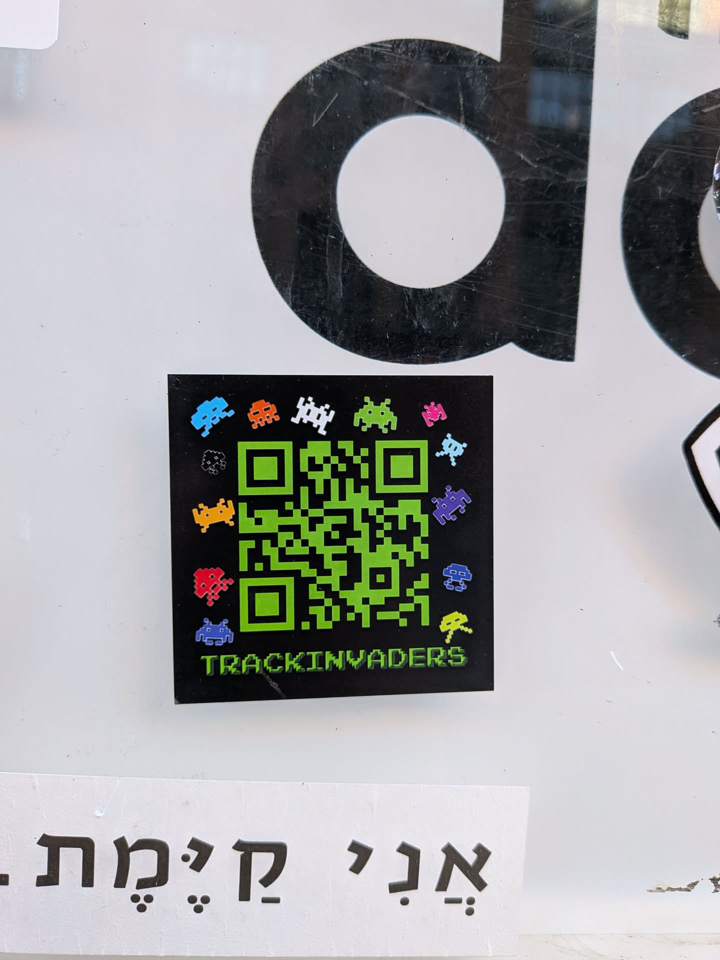 A square-shaped sticker featuring a vibrant green QR code against a black background. Surrounding the QR code are numerous small, pixelated alien characters, reminiscent of the classic Space Invaders game, rendered in a variety of bright colors including blue, orange, white, green, pink, black, yellow, red, and purple. Below the QR code, the word 'TRACKINVADERS' is written in a pixelated green font, matching the QR code's color. The sticker has a glossy finish and is applied to a white surface.