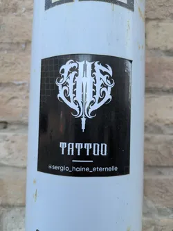 Street sticker The sticker is black with white graphics and text. At the top, there's a stylized emblem resembling wings or decorative flourishes. Below that, the word 'TATTOO' is printed in a bold font. At the bottom, it displays the Instagram handle @sergio_haine_eternelle.