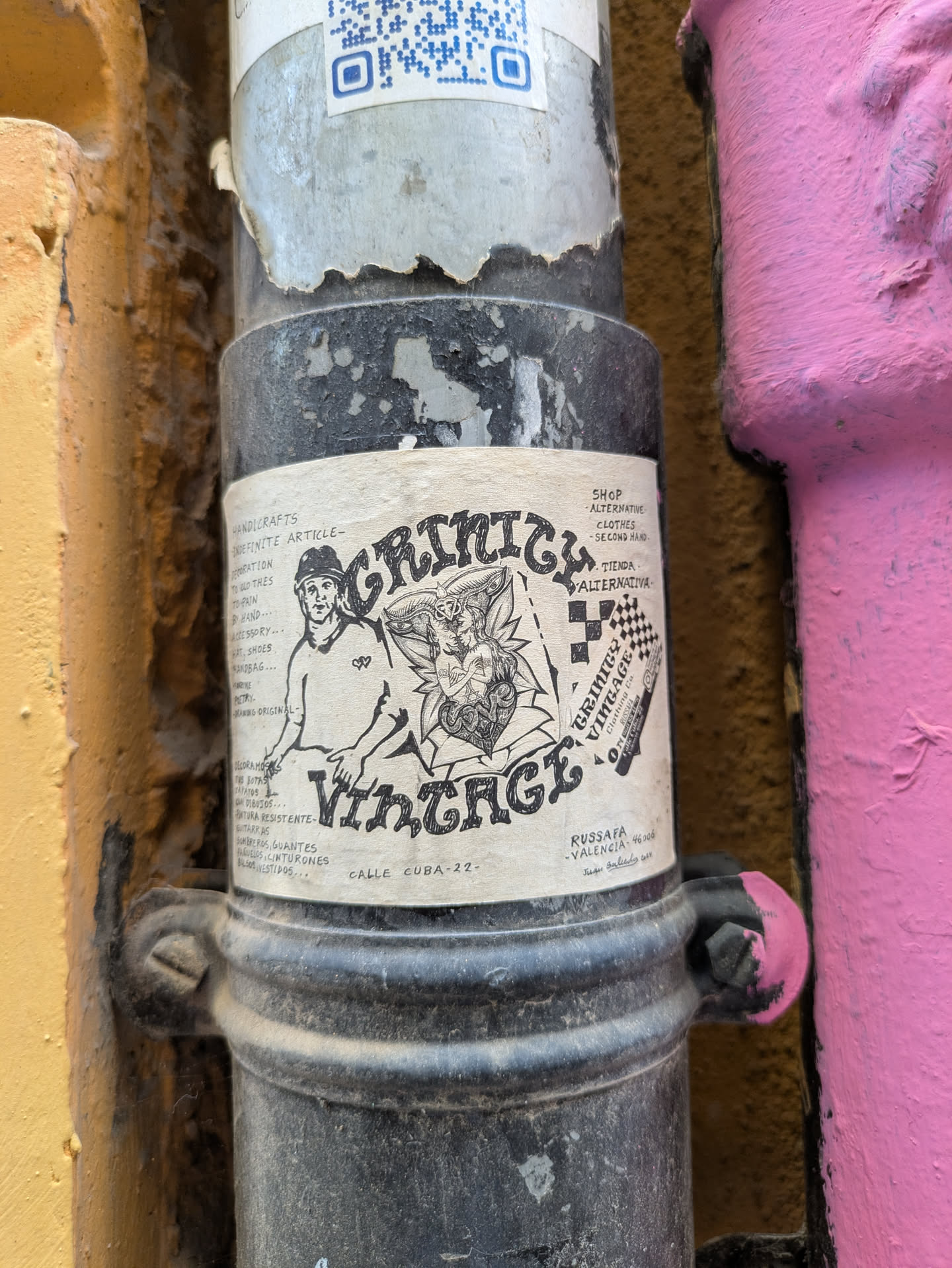 The sticker for Grinicy Vintage Clothing is attached to a black metal pipe. The sticker is white with black ink illustrations and text. It features a man in a hat and a winged figure. Text on the sticker reads 'Grinicy Vintage'. The sticker also mentions the address 'Calle Cuba 22' and 'Russafa-Valencia-46006'.