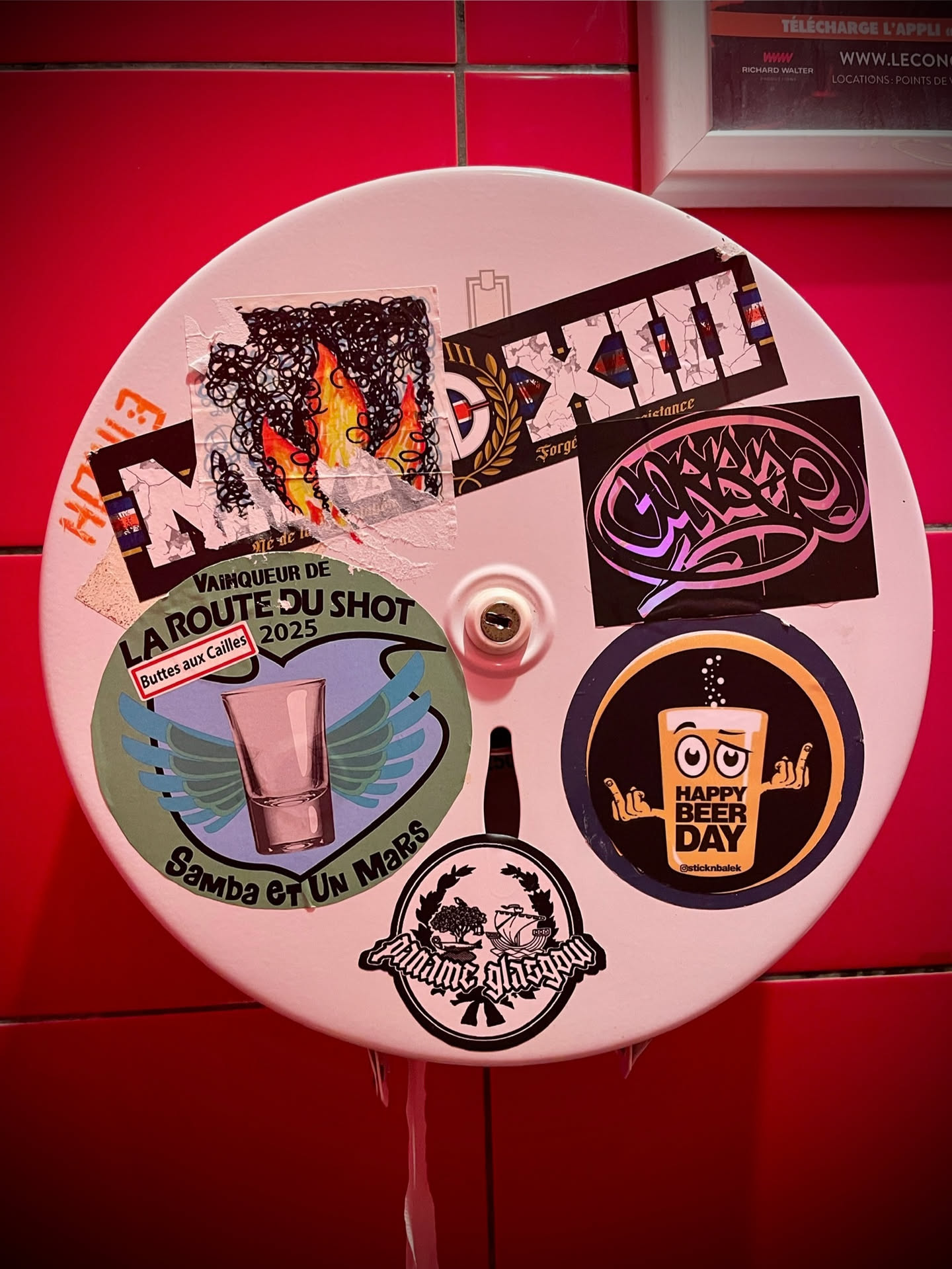 The image centers on a circular, light-colored surface, likely a dispenser, covered with a diverse array of stickers. In the top left, an abstract sticker features black swirling lines over orange and yellow flames. Adjacent to it, a black sticker with white text reads 'MXIII' in a blocky, industrial font, with smaller text 'Forge' and 'Resistance'. Above the center, a stylized graffiti sticker in iridescent pink and purple hues is visible. To the left, a large green and blue circular sticker depicts a shot glass with light blue wings, surrounded by text. Below it, a black and white circular sticker features a crest with trees and a ship, with 'Ancienne Glasgow' in a gothic script. To the right, a black and yellow circular sticker shows a cartoon beer glass with eyes and arms giving two middle fingers, with 'HAPPY BEER DAY' and a social media handle. The stickers show signs of wear, tear, and overlapping, indicating genuine application.