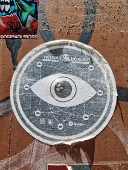 Street sticker Round sticker with an eye symbol in the center, surrounded by a geometric pattern and social media icons. The text "IKIGAI COSMONAUT" is written twice on the top. The sticker has a worn and weathered appearance, showing signs of peeling at the edges.