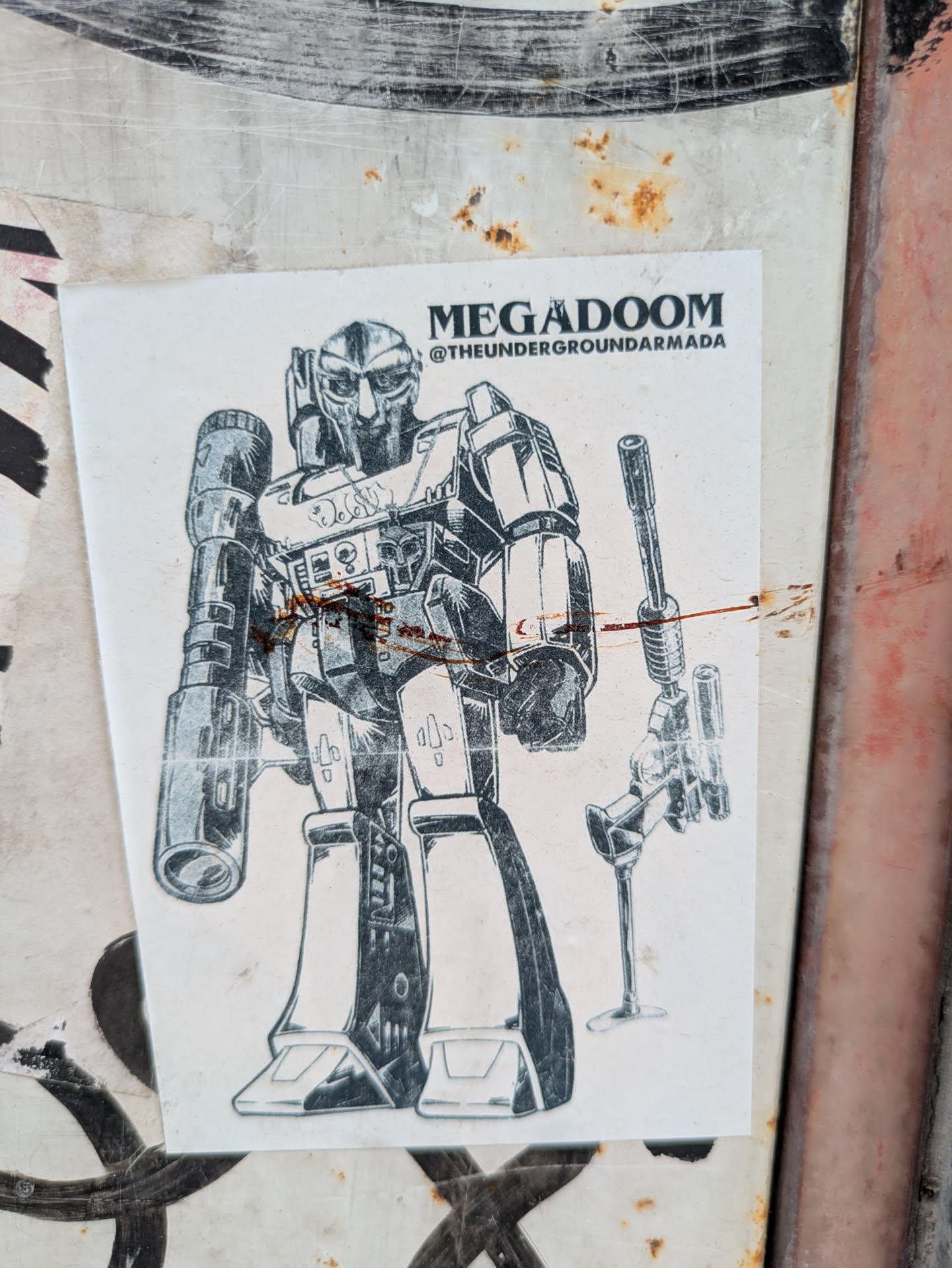The sticker depicts a robot with a gun on its side and the text 'MEGADOOM' and '@THEUNDERGROUNDARMADA' at the top. The sticker is on a weathered surface.