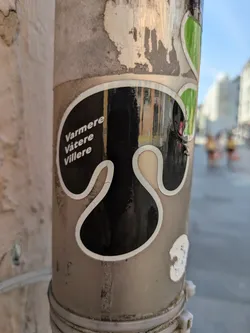 A close-up view of a black sticker with a wavy white outline, affixed to a heavily worn and discolored utility pole. The sticker features three lines of white text, vertically aligned, which read 'Varmere', 'Våtere', and 'Villere'. The pole itself shows signs of age, with peeling remnants of other stickers and a textured, grimy surface. In the background, out of focus, buildings and blurry figures suggest an urban setting under a clear sky.