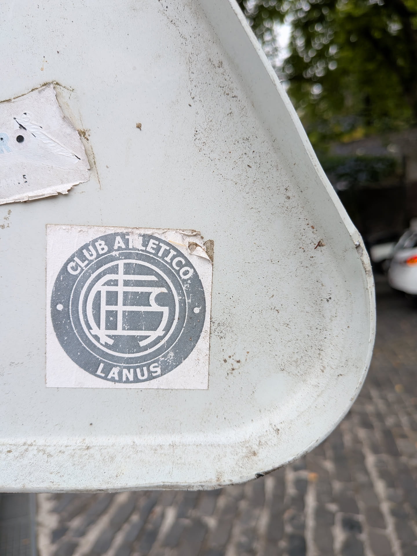 A square sticker with a circular grey emblem. It features the text 'CLUB ATLETICO' and 'LANUS' wrapped around a central stylized monogram on a white background.