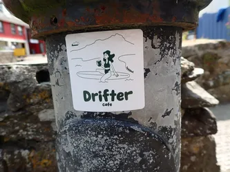 Street sticker Square white sticker with rounded corners. Features dark green line art of a woman in a swimsuit holding a cup while sitting on a surfboard against a coastal mountain backdrop. Below, bold text reads 'Drifter cafe'.