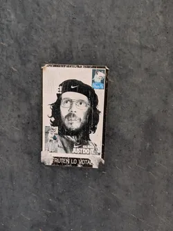 Street sticker The sticker features a monochrome image of a man with a beard and glasses, wearing a beret with the Nike swoosh. Text on the sticker reads "CHE NIKE!" and "KILLINIGO. JUST DO IT."  It also says, 'FRUTEN LO VOTAN'. There is another sticker fragment on the top right that says 'Cheppa Pig'.