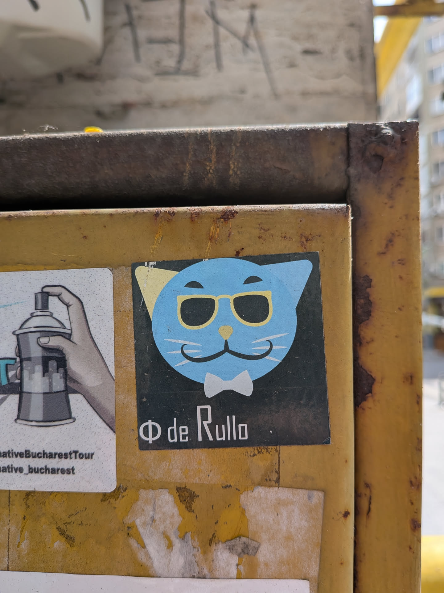 Square black sticker depicting a light blue cat head with yellow sunglasses, a white bow tie, and a mustache-like mouth. Features the text 'Φ de Rullo' at the bottom.
