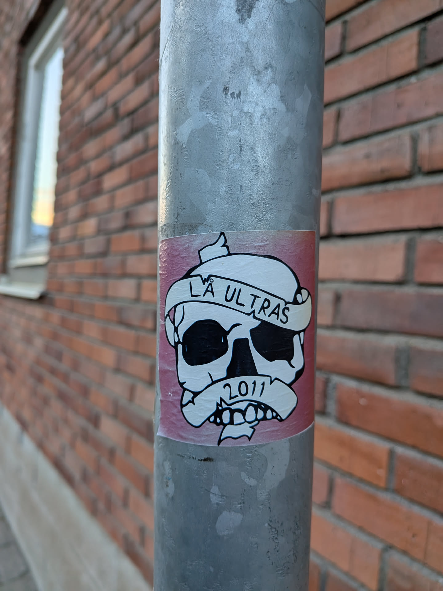 The sticker features a black and white illustration of a skull with large, dark eye sockets and a grinning mouth. A white banderole is wrapped around the skull's forehead, bearing the text 'LÅ ULTRAS' in black, capital letters. Below the skull, another banderole contains the year '2011' in black numerals. The sticker has a pinkish-red background with a white border. The overall style is graphic and somewhat aggressive.