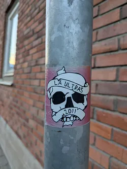 Street sticker The sticker features a black and white illustration of a skull with large, dark eye sockets and a grinning mouth. A white banderole is wrapped around the skull's forehead, bearing the text 'LÅ ULTRAS' in black, capital letters. Below the skull, another banderole contains the year '2011' in black numerals. The sticker has a pinkish-red background with a white border. The overall style is graphic and somewhat aggressive.