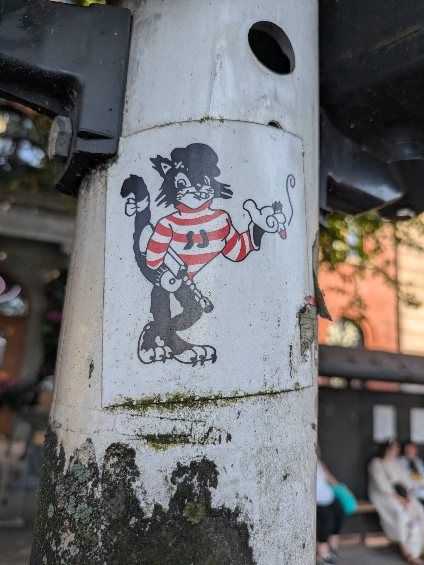 A black and white cartoon cat character is depicted on a sticker applied to a light-colored utility pole. The cat wears a red and white striped long-sleeve shirt with two 'JJ's visible on the chest, a black beret, and appears to be holding a lit cigarette or joint with smoke curling upwards. Its tail is raised and curled, and it has large, stylized feet. The overall style is reminiscent of classic animation or comic characters. The pole itself shows signs of wear and tear, with some dark stains and moss near the base.