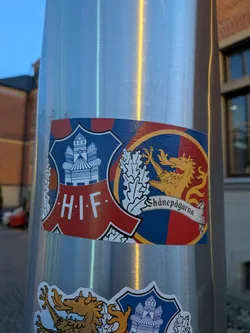 Street sticker The sticker is shield-shaped and divided into two halves. The left half features a blue background with a white depiction of a castle-like structure with multiple towers and arched windows, and the text 'H.I.F.' in white capital letters below it. The right half has a red and blue checkered background with a golden dragon or griffin-like creature in the center, holding a white banner with the text 'Skånepågarna' inscribed on it. The sticker has a red border with white trim.
